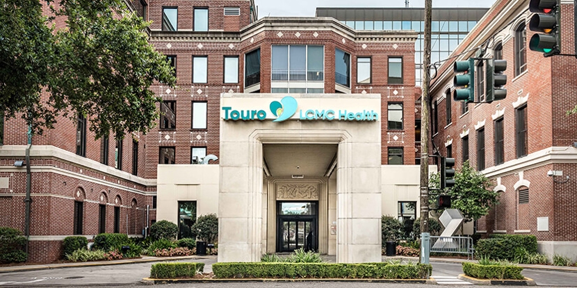 Front entrance of Touro LCMC Health, featuring a stone portico with the “Touro LCMC Health” sign above the entryway. The entrance is centered between multi-story red brick buildings, with landscaped shrubs and plants in the foreground. Traffic lights, trees, and city street elements frame the scene, creating an urban hospital campus setting.