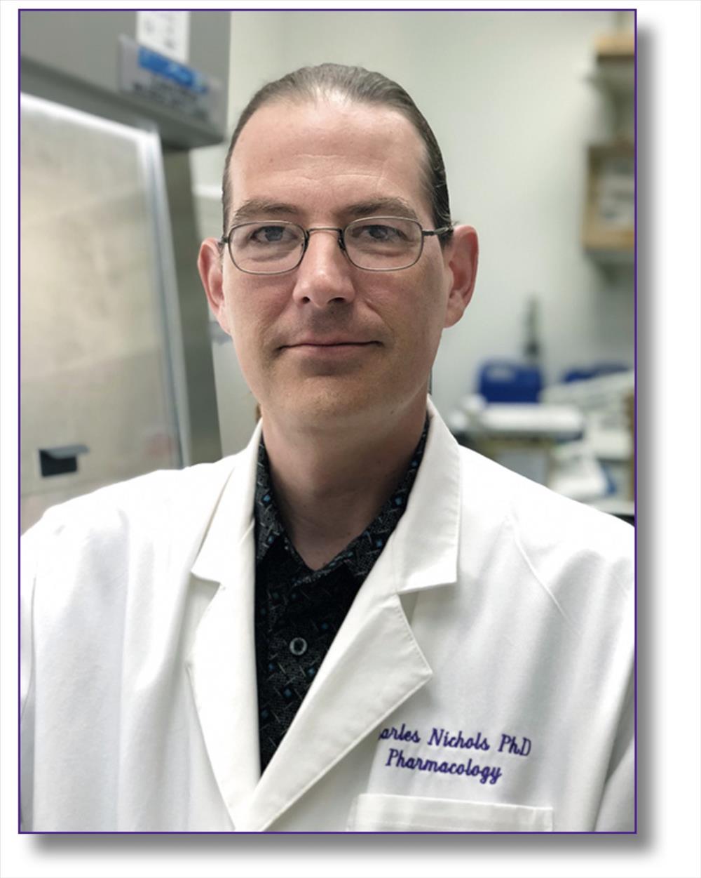 Person wearing thin-framed eyeglasses, a dark collared shirt, and a white laboratory coat, photographed in a laboratory setting. Laboratory equipment and work areas are visible in the softly blurred background. The person is facing the camera, and the image is cropped from the chest up. Blue embroidered text is visible on the lab coat, and the portrait is framed with a thin purple border. The lighting is even, with the laboratory environment providing scientific and professional context.