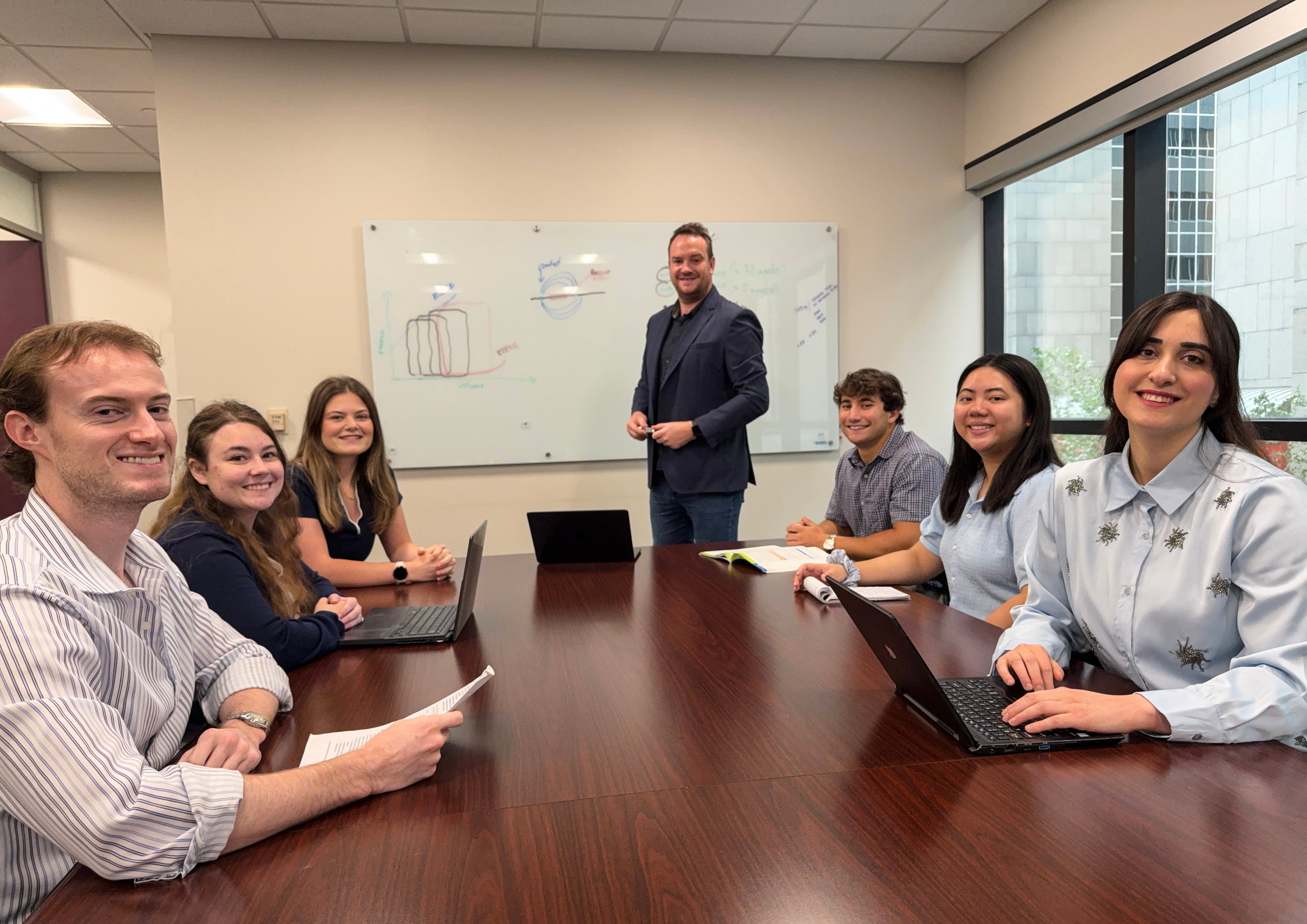 A group of seven people gathered around a conference table in an office meeting room, with one person standing in front of a whiteboard displaying hand-drawn charts and diagrams.