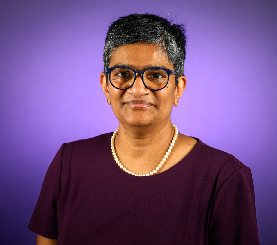 Professional head-and-shoulders portrait of a person with short gray hair and dark blue glasses, wearing a dark purple top and a pearl necklace, posed against a softly lit purple background.