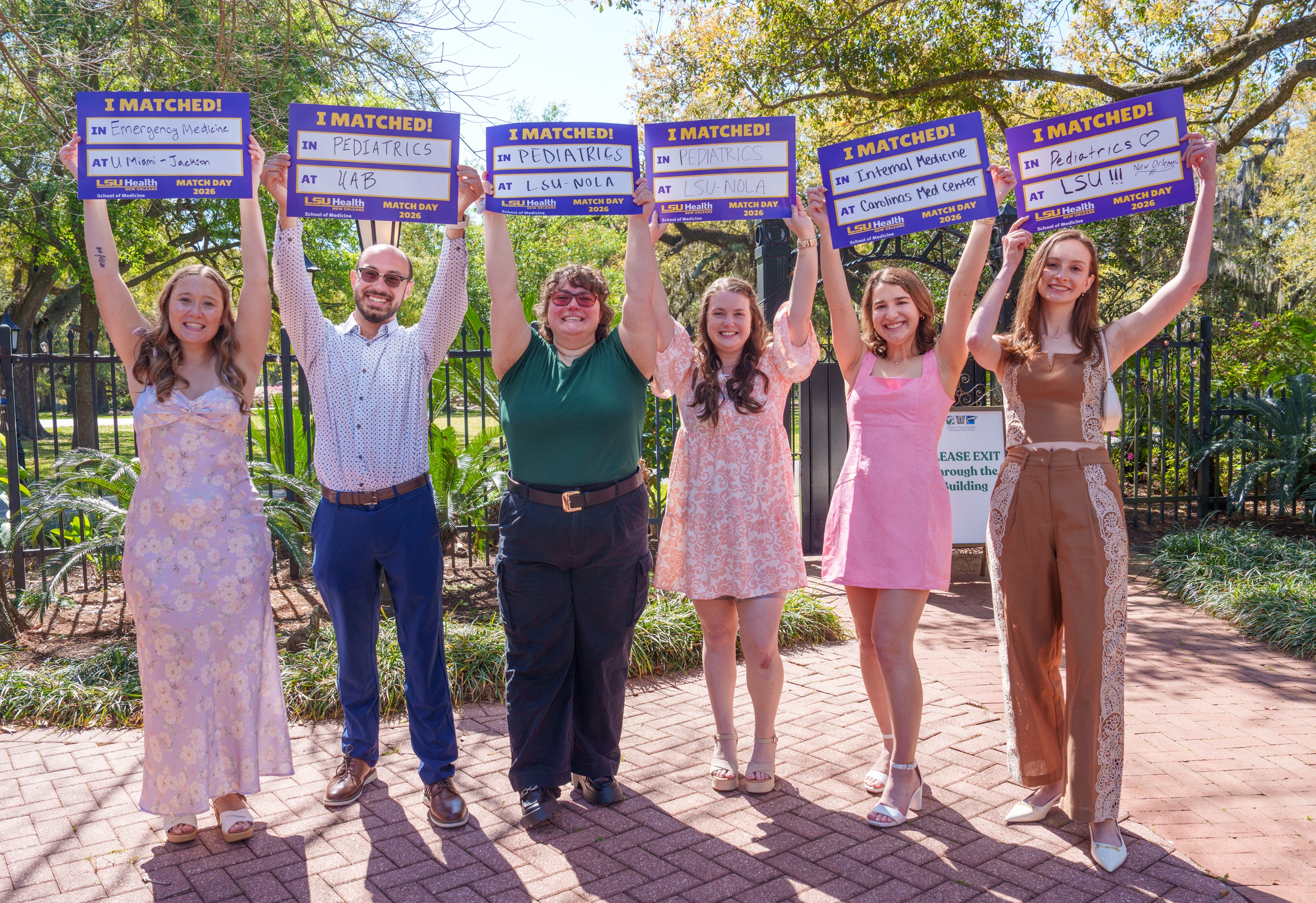 Six people stand on a brick walkway outdoors beneath large trees, each holding an LSU Health School of Medicine “I MATCHED!” sign above their head on Match Day 2026. The signs show residency placements in Emergency Medicine, Pediatrics, and Internal Medicine at institutions including UMiami-Jackson, UAB, LSU-NOLA, Carolinas Medical Center, and LSU. A black iron fence and landscaped greenery are visible in the background on a sunny day.